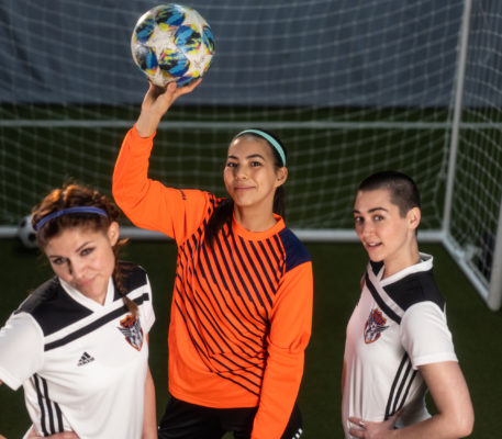 The Wolves actors. Three women in soccer jerseys standing together with the goalie in the middle in an orange shirt, holding a soccer ball, smiling.