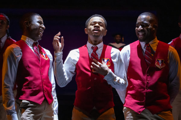 three young black men in red vests, two laughing flanking one of the boys singing